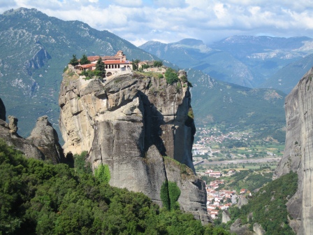 Rock tower with stone monastery on the rop with mountains in the distance and a town below and behind
