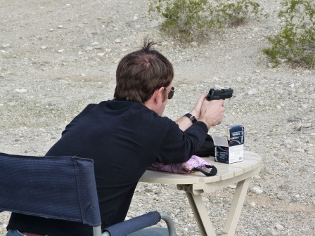 Patrick shooting a handgun seated with arms outstretched on a table