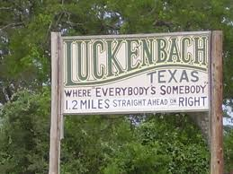 A large white roadside sign saying, "Luckenbach,Texas, Where Everybody's Somebody, 1.2 Miles, Straight Ahead on Right"